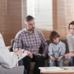 Rear view of female psychologist helping young family with a kid to solve child development problems. Family sitting on a sofa in the blurred background