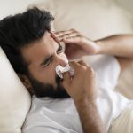 Sick Young Indian Man Blowing Nose In Tissue While Lying In Bed
